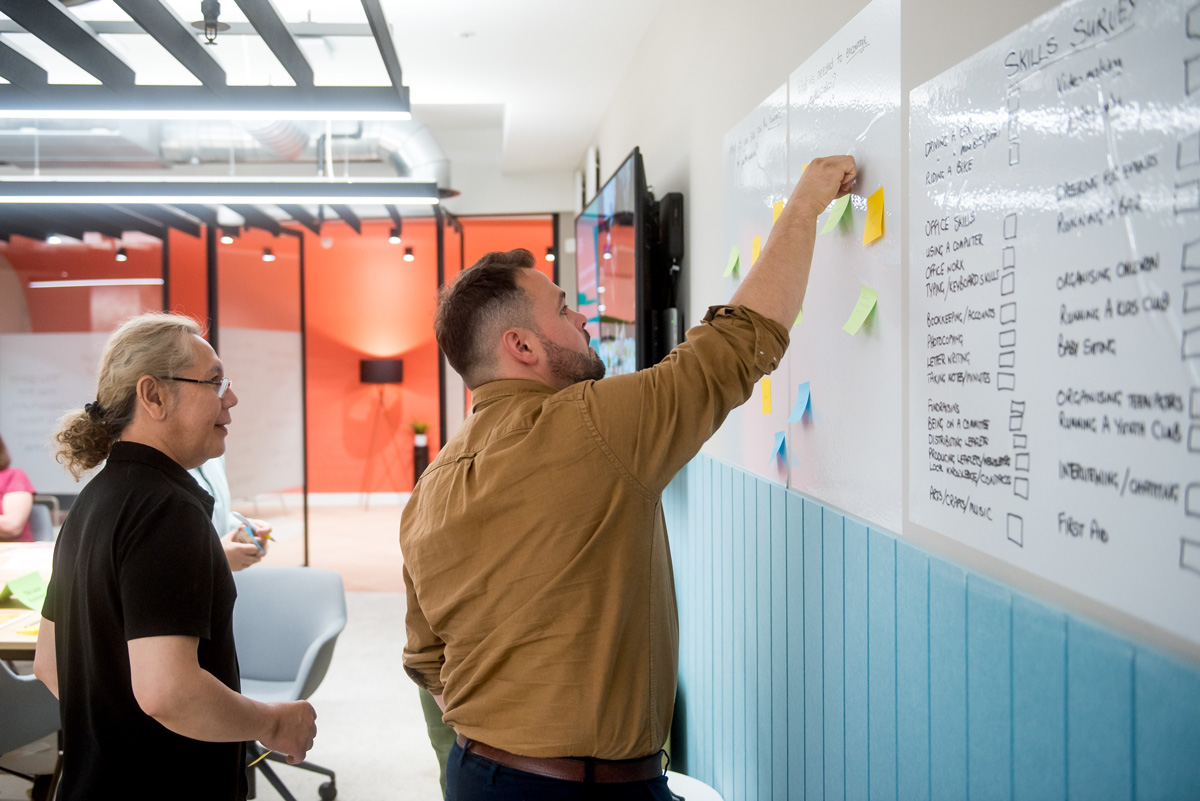 A man is standing at a wall-mounted whiteboard, placing colourful sticky notes in a column next to a list of skills and tasks written on the board. Several notes in blue, green, and yellow are already attached. Another participant stands nearby, observing the activity.