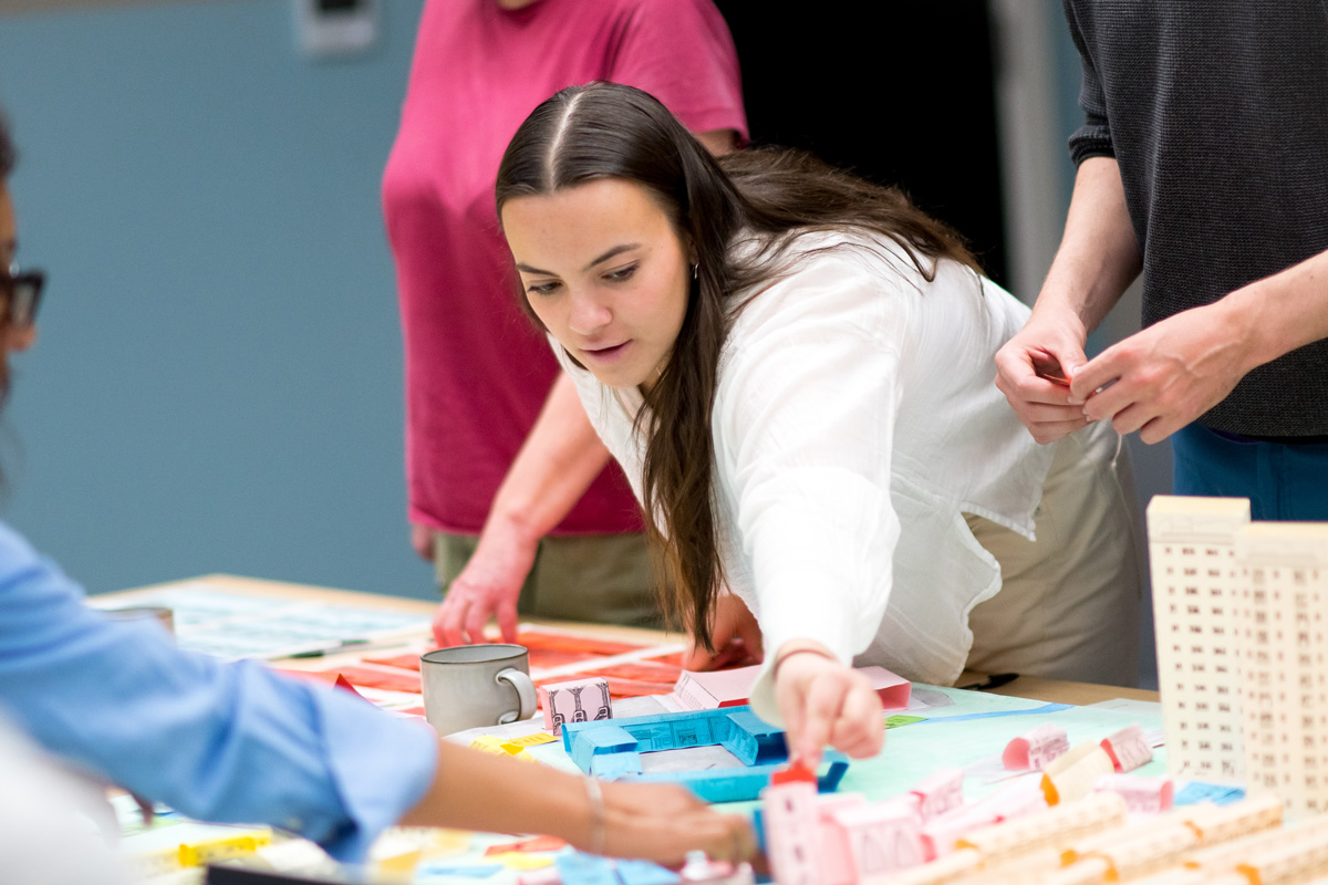 Participants are gathered around a large table, placing small paper and card model buildings onto a colourful map of a neighbourhood. The map includes clearly marked areas and zones, and the models represent different types of structures in various colours such as blue, pink, and beige.