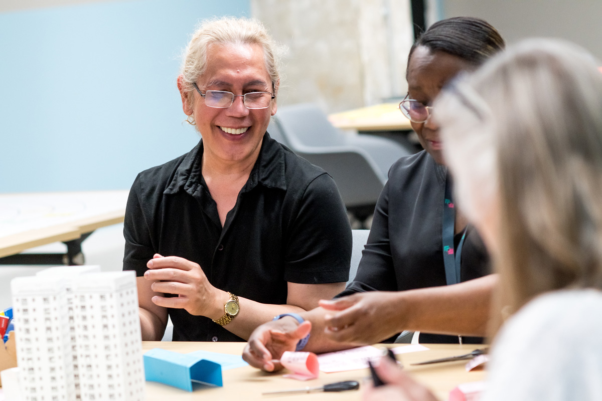 Participants sit around a table constructing a scale model of a neighbourhood during a Planning for Real training session. On the table are small paper and card structures, including a white multi-storey building model and blue and pink folded pieces representing other buildings or features. Pens and other craft materials are scattered across the workspace, indicating an interactive, hands-on planning activity.