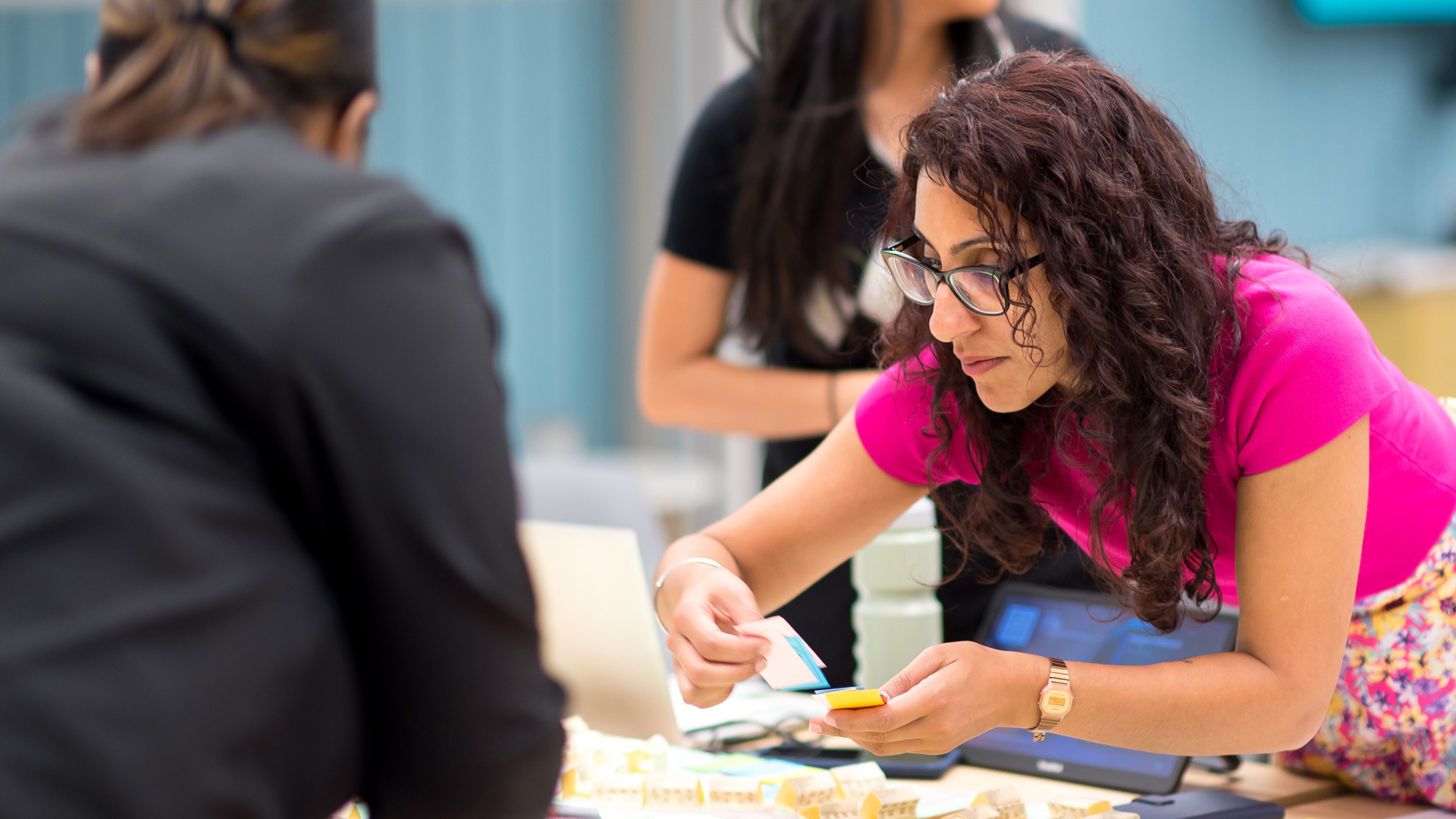A woman in a bright pink top is leaning over a table, sorting through small rectangular cards. The table is covered with various materials, including a tablet device and other cards. Another participant is stood behind, engaged in the activity.